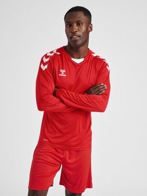 Homme debout, les bras croisés, portant un maillot de sport rouge à manches longues et un short assorti sur un fond uni.