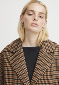 Smeđi i crni kaput s uzorkom houndstooth s urezanim ovratnikom, izrađen od teksturirane tkanine. Vidljive srebrne kružne naušnice. Crni top ispod.
