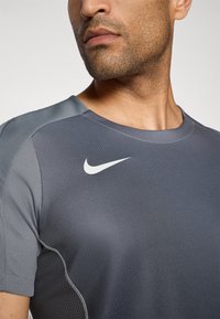 Grå idretts skjorte med rund hals. Har teksturert stoff, kontrasterende skulderpaneler og en hvit Nike-logo på brystet.