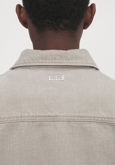 Lichtgrijze denimjack met kraag, met een klein logo op de achterkant van de nek. De stof heeft een gladde, gestructureerde afwerking.
