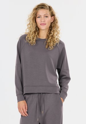 Jonge vrouw met krullend blond haar draagt een losse grijze trui en bijpassende broek met trekkoord, staand met één hand in de zak.