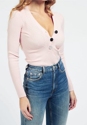 Frau trägt ein hellrosa, geripptes Langarmshirt mit drei großen Knöpfen und hochtaillierte blaue Jeans, steht mit entspannten Armen.