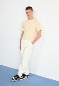 Beige bomulds t-shirt med præget logo, hvide bukser og sorte sneakers med mønstrede accenter. Modellen står foran en hvid væg.