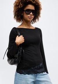 Γυναίκα με σγουρά μαλλιά που φοράει μαύρο μακρυμάνικο τοπ με δαντελωτό τελείωμα, υπερμεγέθη μαύρα γυαλιά ηλίου, ανοιχτό μπλε τζιν και κρατάει μαύρη δερμάτινη τσάντα ώμου.