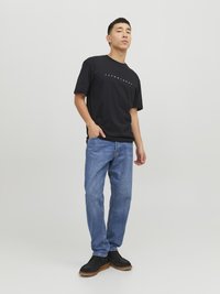 Schwarzes T-Shirt mit kurzen Ärmeln und weißem Logo, kombiniert mit blauen Denim-Jeans. Das Modell trägt schwarze Schuhe mit einer strukturierten Sohle.