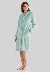 Mujer de pie descalza en una bata de baño de textura ligera y azul claro, de largo hasta las rodillas, con cuello y bolsillos delanteros, mirando hacia un lado sobre un fondo liso.