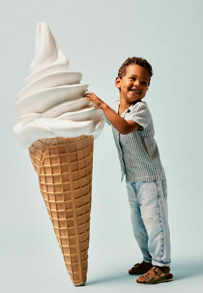 Un enorme cono gelato con una soffice crema bianca sulla cima di un cono di cialda beige e testurizzato. Un bambino in una maglietta a righe interagisce con il cono.