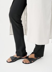 Sandales noires à design à brides croisées, semelles plates et orteils découverts. Portées avec un pantalon noir et une tunique longue de couleur claire.