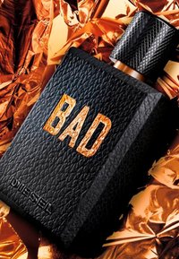 Schwarze, strukturierte Flasche mit dem in Orange geprägten Schriftzug 'BAD'. Verfügt über einen geriffelten schwarzen Verschluss. Der Hintergrund besteht aus zerknittertem metallischem Goldfolien.