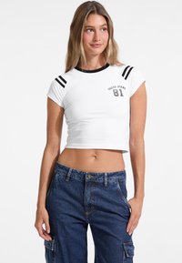 Balta trumpa marškinėliai su juodu apvaliu iškirpimu ir dryžuotomis petnešėlėmis. Ant jų esantis "Guess Jeans 81" logotipas yra pilkas. Modelis dėvi mėlynas cargo kelnes.