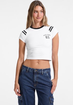 Biela skrátená tričko s čiernym okrúhlym výstrihom a pruhovanými detailmi na pleciach. S logom "Guess Jeans 81" v sivej farbe. Model má na sebe modré cargo nohavice.
