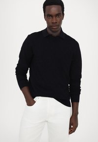 Pull noir anthracite avec un motif diamant texturé, col rond et poignets côtelés, porté par-dessus une chemise noire à col et associé à un pantalon blanc.