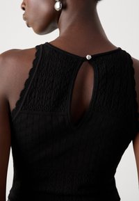 Top in maglia nera con un motivo testurizzato, spalle scallopate e chiusura a chiave sulla schiena con un bottone decorativo.