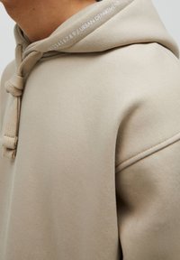 Hoodie beige avec une finition texturée, doté d'un cordon de serrage noué et d'un texte imprimé sur le col. Détails de coutures visibles le long de l'épaule.