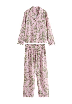 Frau mit langen dunklen Haaren, die einen rosa Blumen-Pyjama trägt und barfuß in einem sanft getönten Wohnzimmer mit pinkem Sofa und heller Dekoration steht.