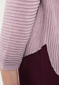 Top de manga larga rosa claro con ribetes y un dobladillo ligeramente asimétrico, combinado con pantalones burdeos profundos. La textura parece suave y acogedora.