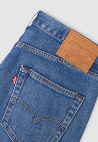 Μπλε τζιν Levi's 501 διπλωμένα, με εμφανή την πίσω τσέπη, την κόκκινη ετικέτα Levi's και το δερμάτινο ταμπελάκι της μάρκας σε ταμπά χρώμα με μέγεθος W32 L.