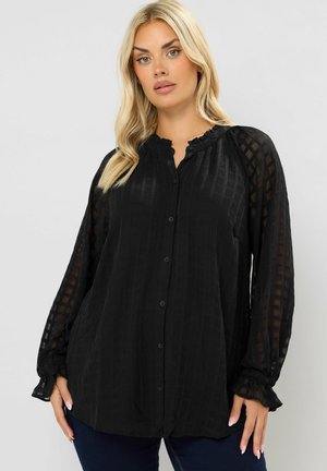 Donna bionda che indossa una blusa nera trasparente a quadri con colletto e polsini arricciati, in piedi contro uno sfondo chiaro e semplice.