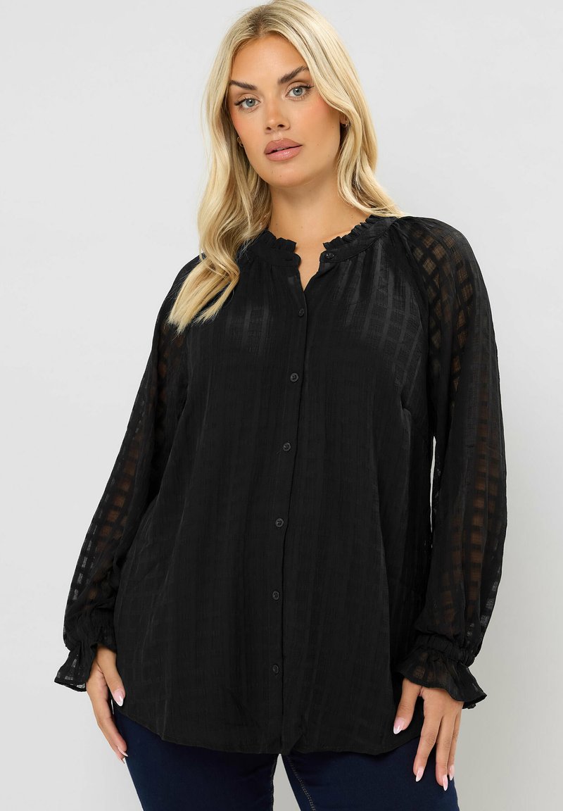 Blonde Frau, die eine schwarze, durchsichtige, kariert gemusterte Bluse mit Rüschensaum und -manschetten trägt, steht vor einem einfarbigen, hellen Hintergrund.