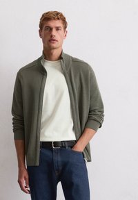 Olijfgroene zip-up cardigan met geribde manchetten en kraag, gecombineerd met een romige ronde hals T-shirt en donkerblauwe jeans, gedragen met een zwarte riem.