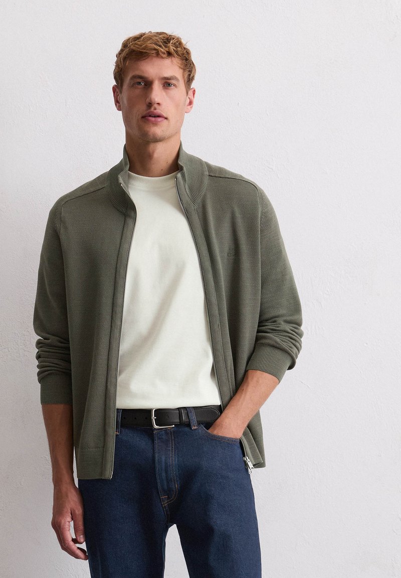 Olijfgroene zip-up cardigan met geribde manchetten en kraag, gecombineerd met een romige ronde hals T-shirt en donkerblauwe jeans, gedragen met een zwarte riem.