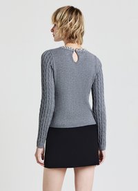 Maglione grigio a maglia con maniche a trecce, dettaglio con apertura a chiave sul retro e decorazione in perle sul collo. Abbinato a una gonna nera.