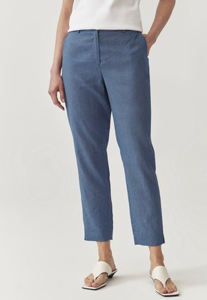 Vrouw draagt blauwe gestructureerde enkellange broek, wit mouwloos topje en witte open-toe sandalen met hak, staat met één hand in de zak.