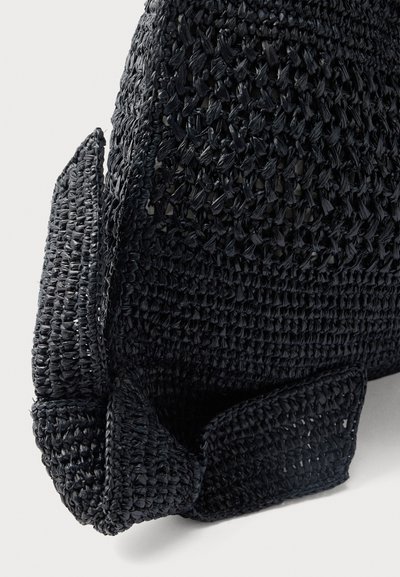 Κοντινό πλάνο μιας μαύρης τσάντας από πλεκτό ραφίας που αναδεικνύει την υφή, τις ραφές και τη λεπτομέρεια με φιόγκο σε ουδέτερο φόντο.