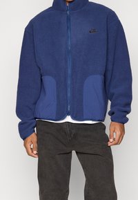 Blå fleecejacka med hög krage, dragkedja fram och två sidofickor. Strukturerad tyg, kontrasterande blå panelaccenter och liten logotyp på bröstet.