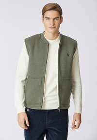 Gilet verde senza maniche con colletto alto, zip frontale e due tasche laterali, indossato sopra un maglione leggero, abbinato a pantaloni scuri.