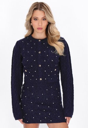 Meriharmaa neulemekko pitkilla hihoilla, pyöreällä kaula-aukolla, jossa on helmimäisiä koristuksia ja metalliset nappikiinnitykset edessä.