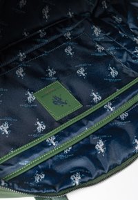 Granatowa podszewka z powtarzającymi się jasnoniebieskimi logo U.S. Polo Assn., z zieloną skórzaną metką na krawędzi i kontrastującym zielonym przeszyciem.
