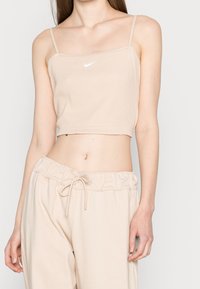Beige vypasovaný crop top s tenkými ramínky a bílým logem Nike uprostřed, v kombinaci s odpovídajícími běžnými béžovými kalhotami.