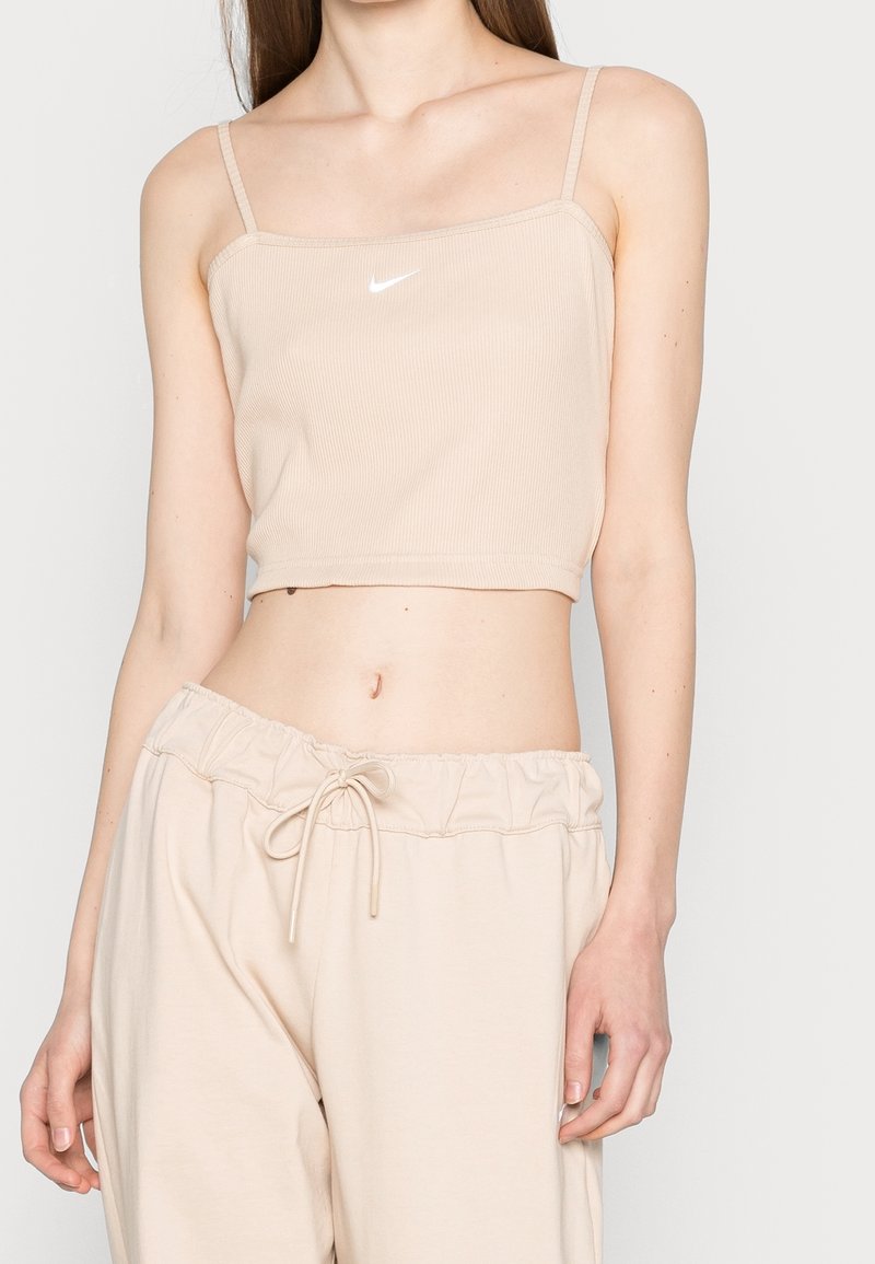 Beige vypasovaný crop top s tenkými ramínky a bílým logem Nike uprostřed, v kombinaci s odpovídajícími běžnými béžovými kalhotami.