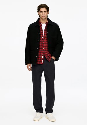 Junger Mann in einer schwarzen Knopfjacke über einem rotkarierten Hemd, schwarzen Hosen und weißen Sneakers, der vor einem weißen Hintergrund steht.