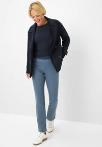 Cappotto doppiopetto blu navy su un top blu navy a coste, abbinato a pantaloni aderenti azzurri e sneakers bianche. Texture lisce e vestibilità sartoriale.