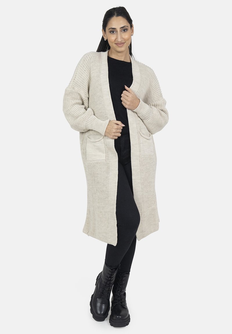 Cárdigan tejido beige con mangas largas, apertura frontal y dos bolsillos frontales, combinado con un atuendo negro y botas negras resistentes.