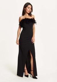 Black, off-the-shoulder dress with feather accents at the neckline, fitted bodice, and side slit. Paired with black high-heeled shoes.