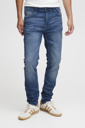 Un mannequin porte un jean bleu slim, une chemise blanche et des baskets blanches et beige sur un fond uni.