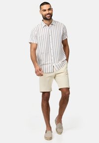Chemise à manches courtes et boutons, avec des rayures verticales dans des couleurs pastel. Associée à un short beige et des chaussures à enfiler de couleur claire.