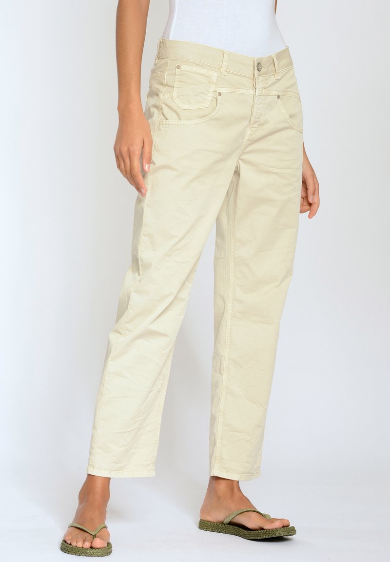 Pantalon beige slim porté par une personne debout avec des tongs vertes et un haut blanc rentré, sur un fond uni.