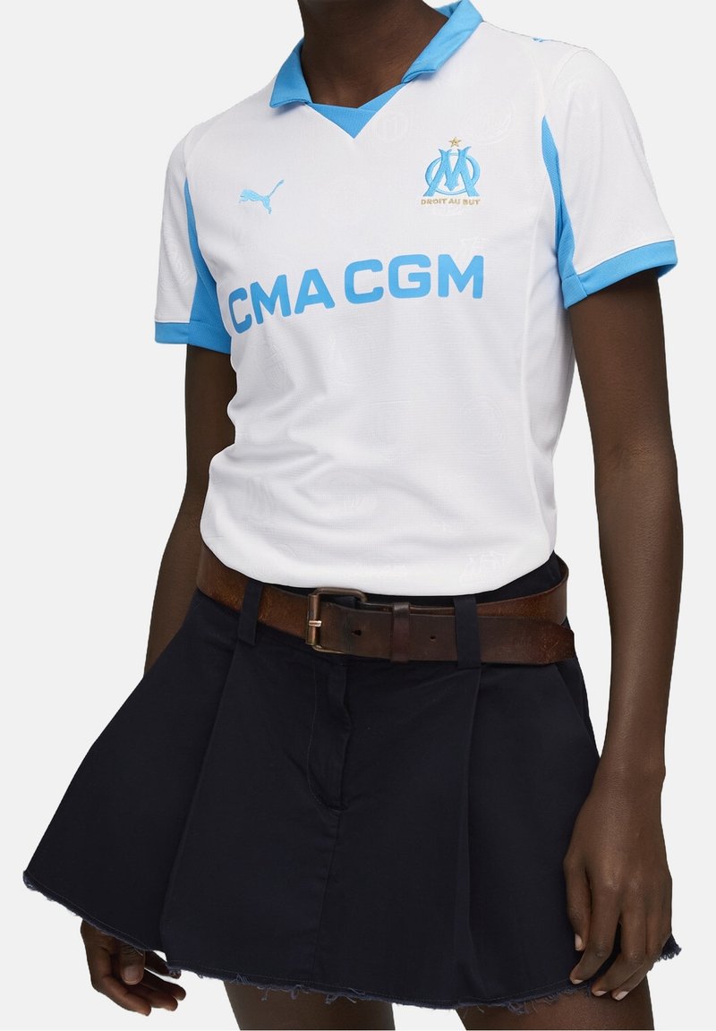 Osoba v bílém a modrém sportovním dresu Puma Marseille, uvrženém do černé sukně, s hnědým páskem.