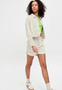Giacca cropped a righe beige e pantaloni shorts abbinati, abbinati a una maglia verde. Sneakers con accenti multicolori. Tessuto liscio e leggero.