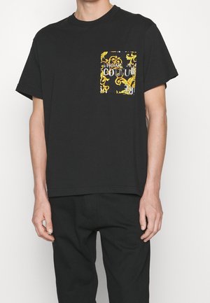 Mężczyzna ubrany w czarny t-shirt z krótkimi rękawami i kieszenią na piersi, zdobiony złotym barokowym wzorem oraz białym napisem „Versace Jeans Couture”.
