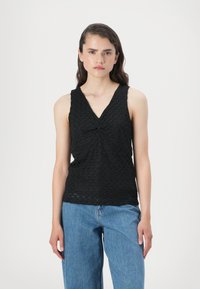 Top preto sem mangas com decote em V e detalhe frontal torcido, apresentando um padrão texturizado, combinado com jeans de perna larga azul claro.