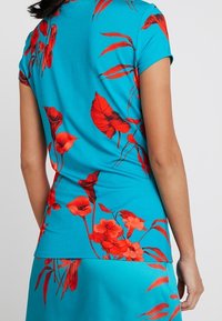 Top de manga corta de color turquesa con patrones florales rojos; textura suave, diseño ajustado y un escote redondeado, combinado con una falda a juego.
