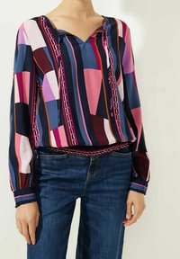 Woman wearing a multicolored geometric patterned blouse with long sleeves paired with blue denim jeans.