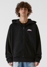 Svart zip-up hettegenser med stor lomme på fronten, snor i hetten og en liten rosa logo på brystet. Mykt stoff med avslappet passform.