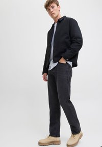 Chaqueta bomber negra con cremallera al frente, camiseta blanca debajo, pantalones oscuros y calzado de cordones en tono marrón claro. Tejido suave y colores neutros.