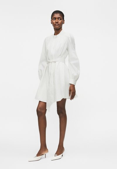 Γυναίκα στέκεται φορώντας λευκό μακρυμάνικο φόρεμα με σφικτή μέση και λευκά μυτερά mules με τακούνι, σε απλό λευκό φόντο.